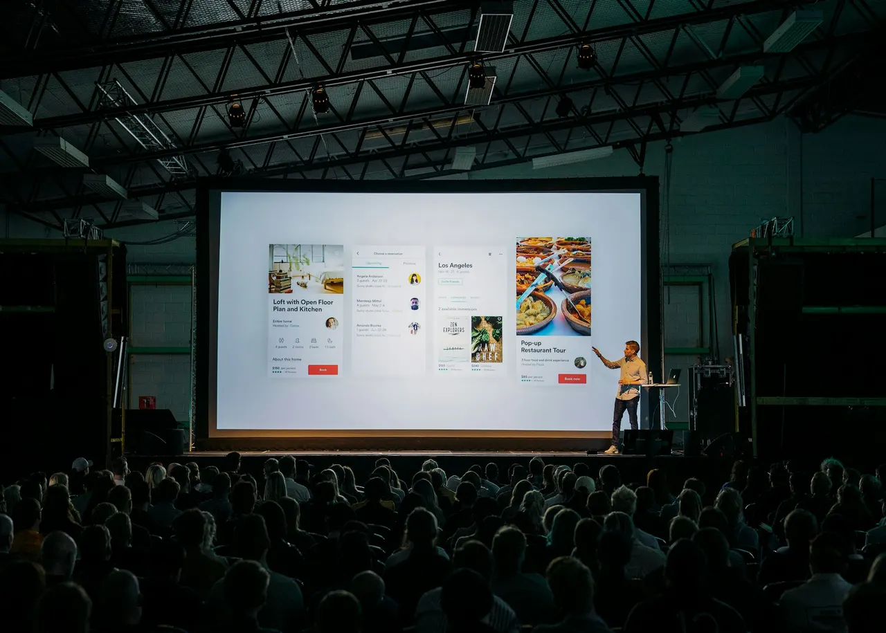 Presenter on stage beside a large projection screen at a corporate general session